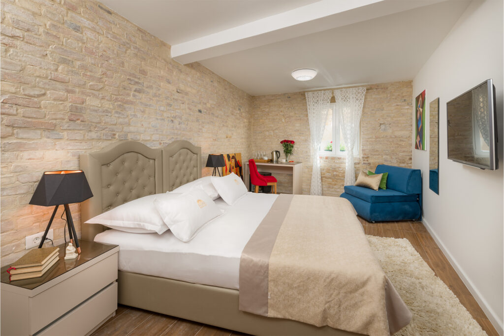 Bright triple room featuring a king‑size bed against brick wall, blue sofa, TV on wall, and partial view of bedside table and lamp.